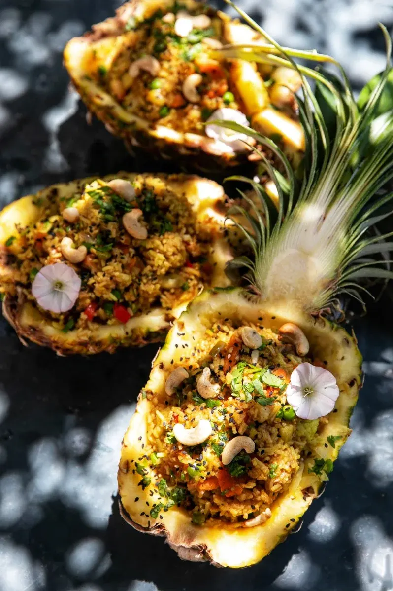 Tre halvor av ananas fyllda med kryddat ris, grönsaker och cashewnötter, garnerade med örter och små blommor.