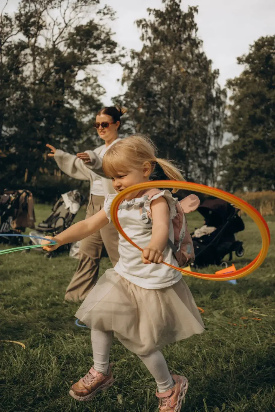 Ett barn leker med färgglada ringar på en gräsplan medan en vuxen står bakom, omgiven av träd och barnvagnar.