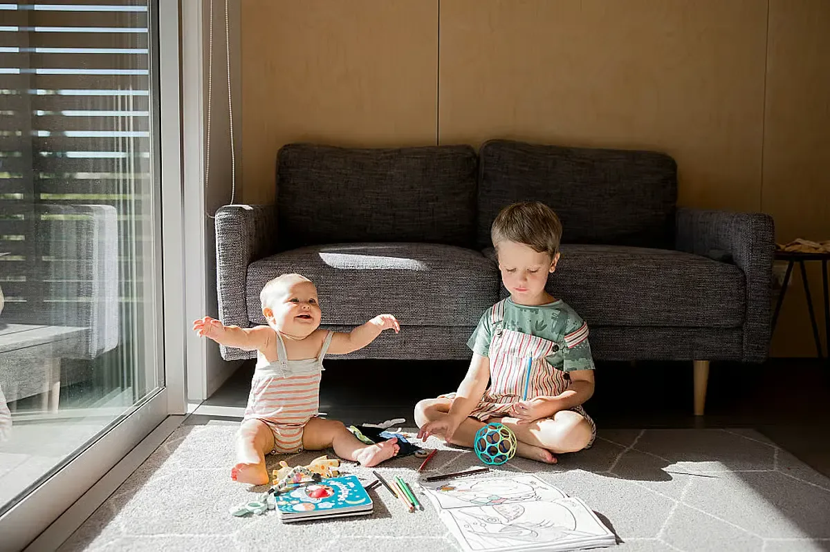 Ett spädbarn och ett äldre barn sitter på golvet framför en soffa och leker med kritor, böcker och leksaker i solljuset.