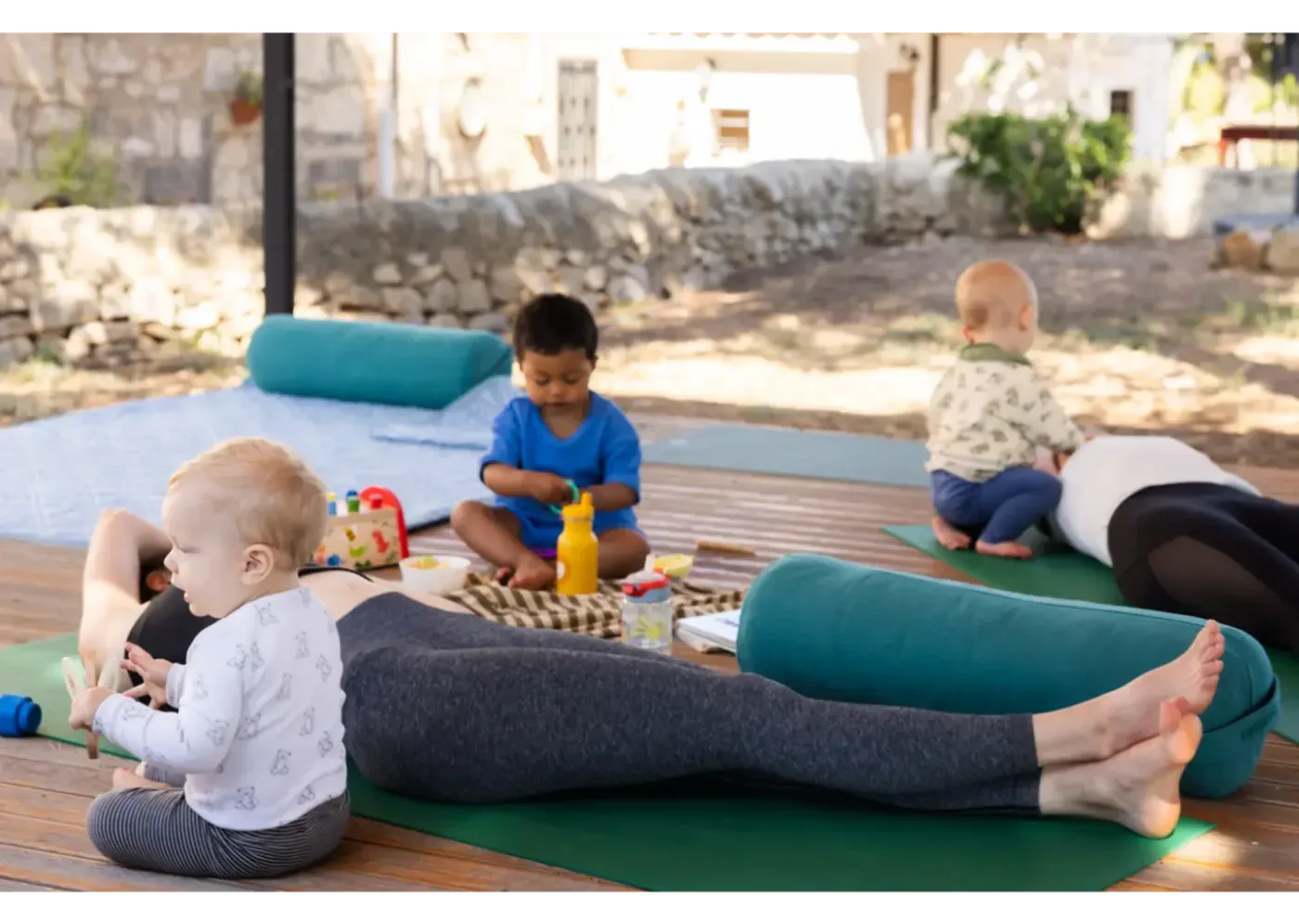 Barn leker på filtar och yogamattor medan en vuxen ligger utsträckt på en matta i en solig innergård.