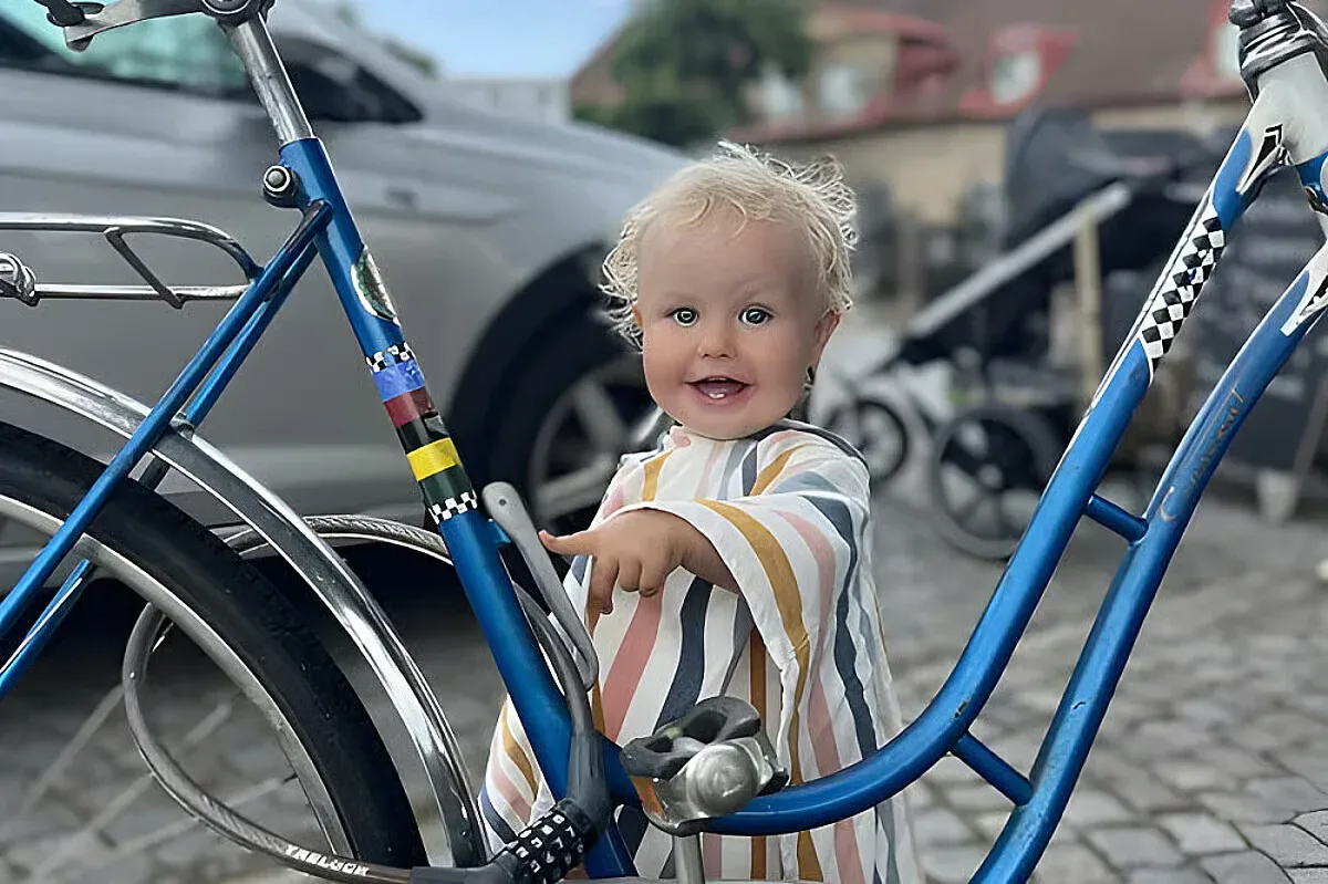 Ett litet barn i randig klänning står mellan två blå cyklar på en kullerstensbelagd gård.