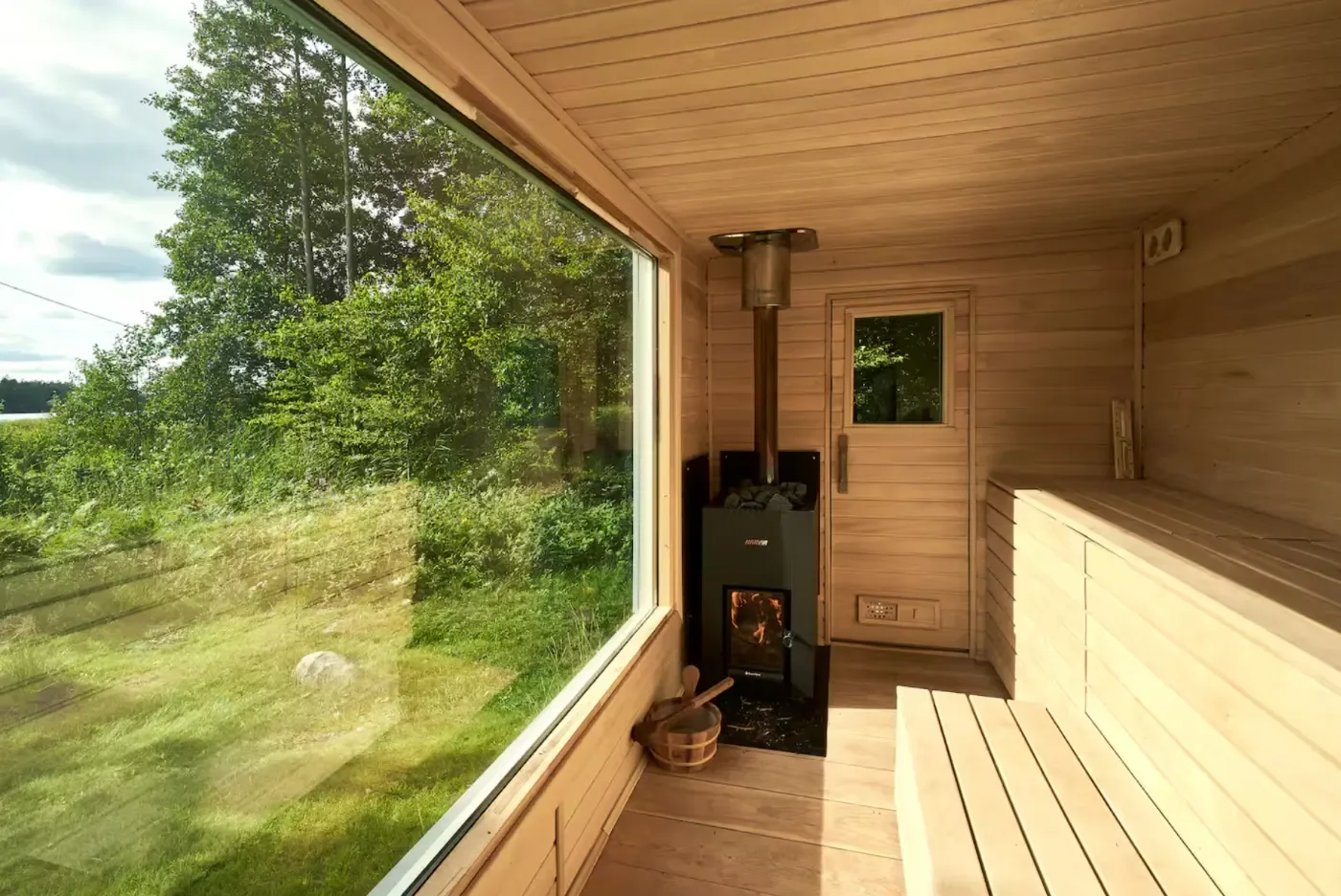 Ljust träbeklätt basturum med vedeldad kamin, lavar och stort fönster med utsikt över grön natur.
