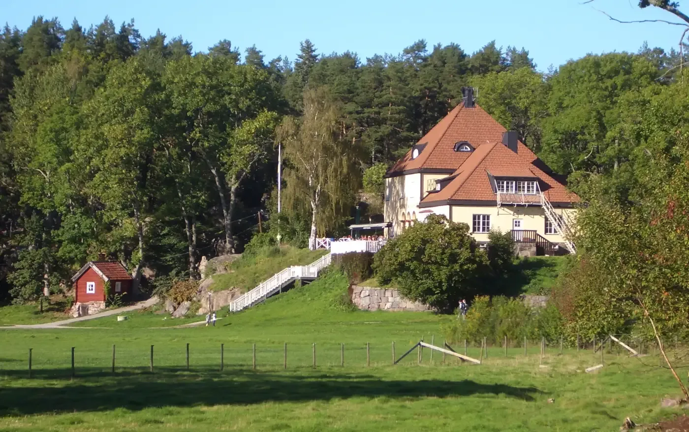 En stor ljus byggnad med röda tegeltak ligger i en grön sluttning bland träd, med en liten röd stuga och gräsplan i förgrunden.