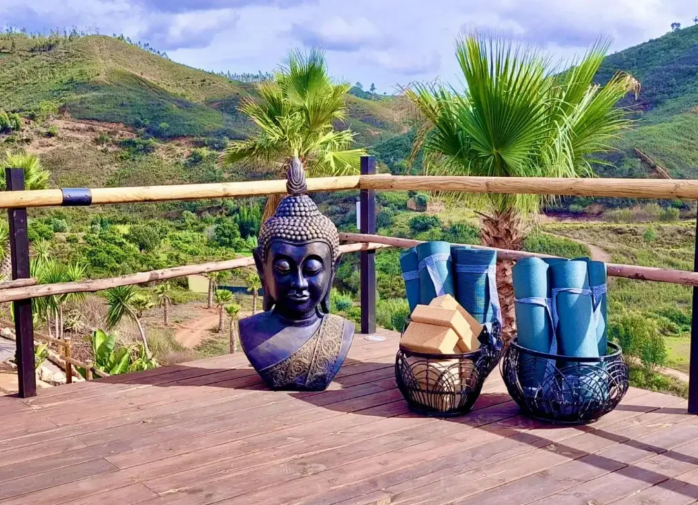 En Buddha-byst, hoprullade yogamattor och träblock står på en träaltan med utsikt över palmer och gröna berg i Algarve.