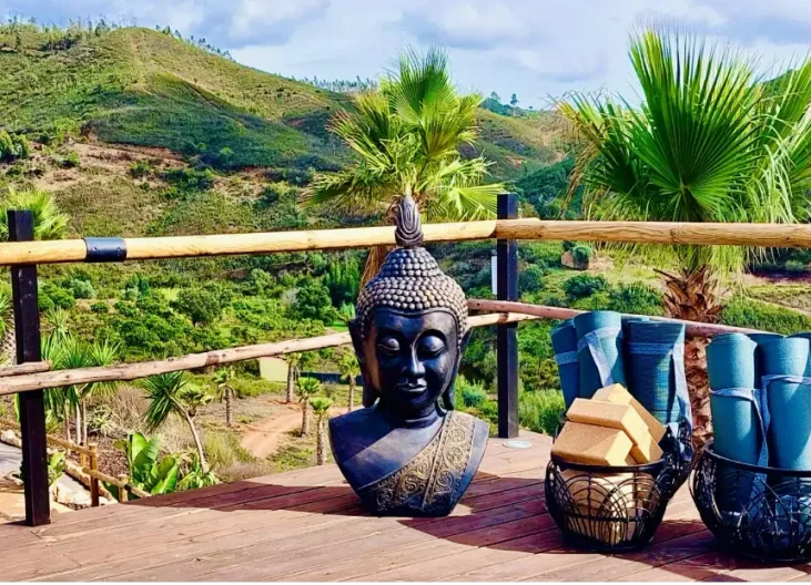 En Buddhahuvudstaty står på en träterrass med yogamattor och kuddar, omgiven av palmer och gröna kullar i Algarve.