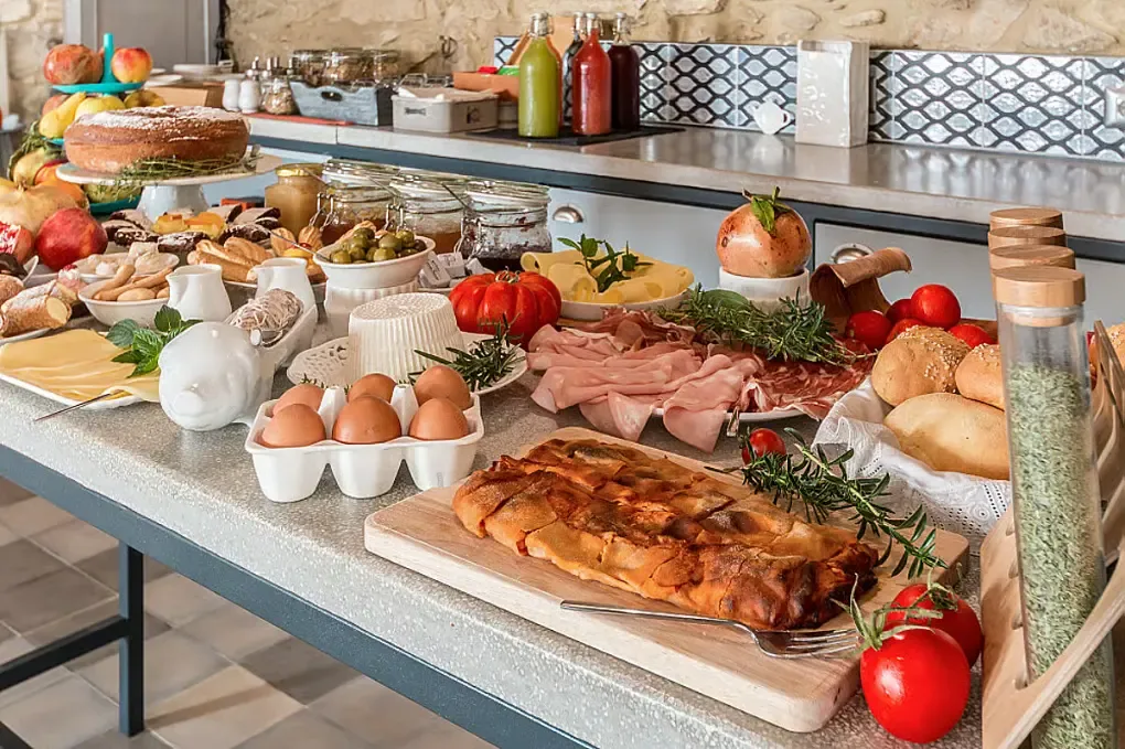 Ett buffébord med bröd, ägg, ostar, charkuterier, grönsaker, frukt och flaskor med dryck i ett lantligt kök.