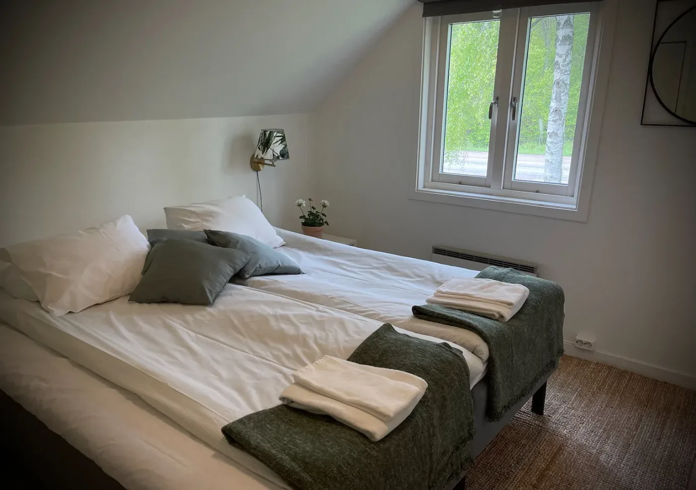 Delat rum med en bäddad dubbelsäng, gröna kuddar, fotpallar med handdukar och ett fönster med utsikt mot björkträd.