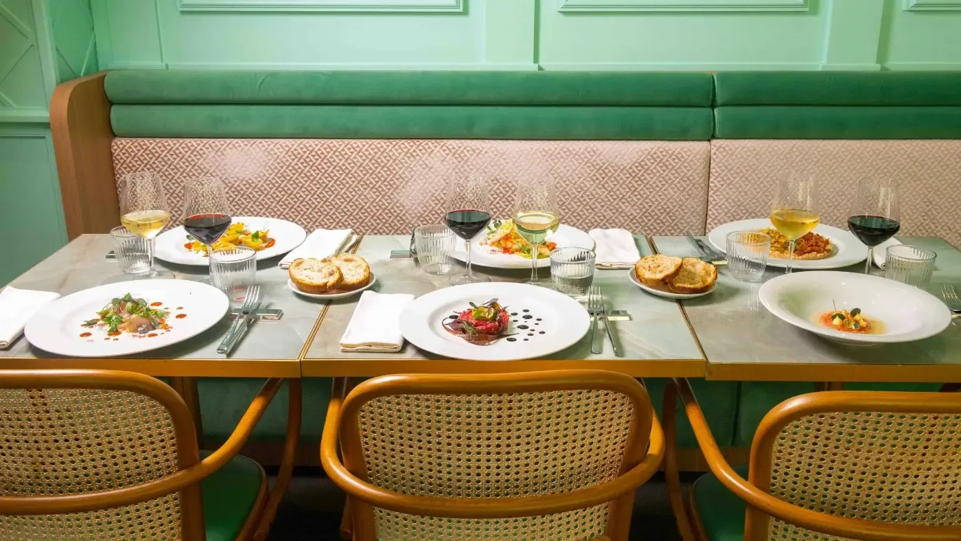 Ett dukat restaurangbord med flera konstfullt upplagda rätter, bröd och vinglas framför en grön sittbänk.