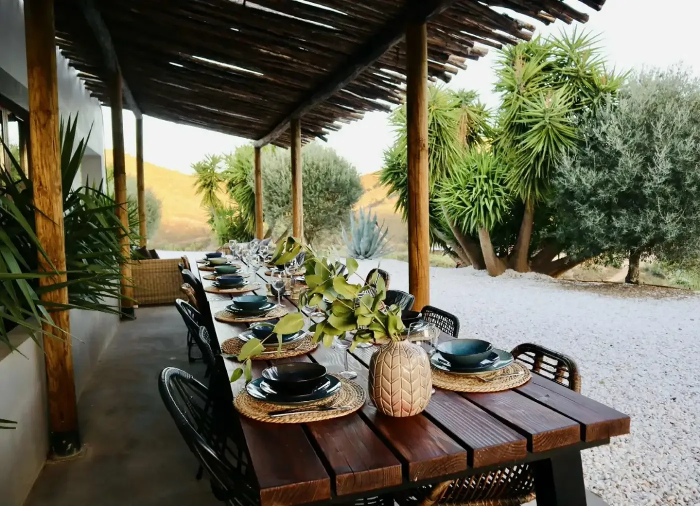 Ett dukat långbord står under en pergola omgiven av grönska, med kullar i bakgrunden.