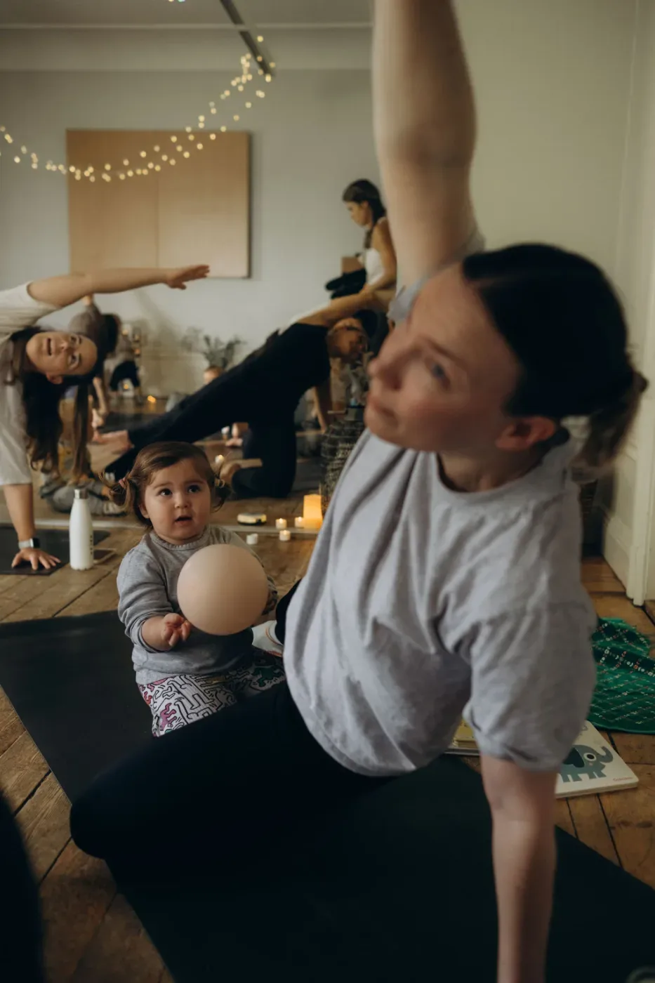 En vuxen och ett barn sitter på golvet medan flera vuxna deltar i en lugn rörelseövning i ett varmt inomhusrum.