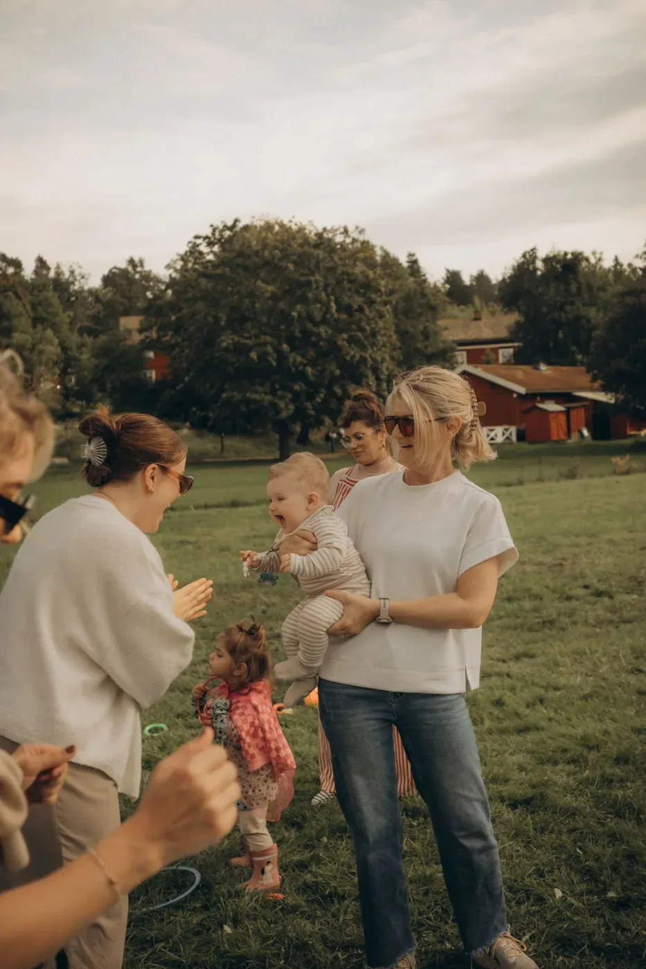 Vuxna och barn samlas på en gräsmatta framför röda stugor, medan en kvinna håller ett litet barn i famnen.