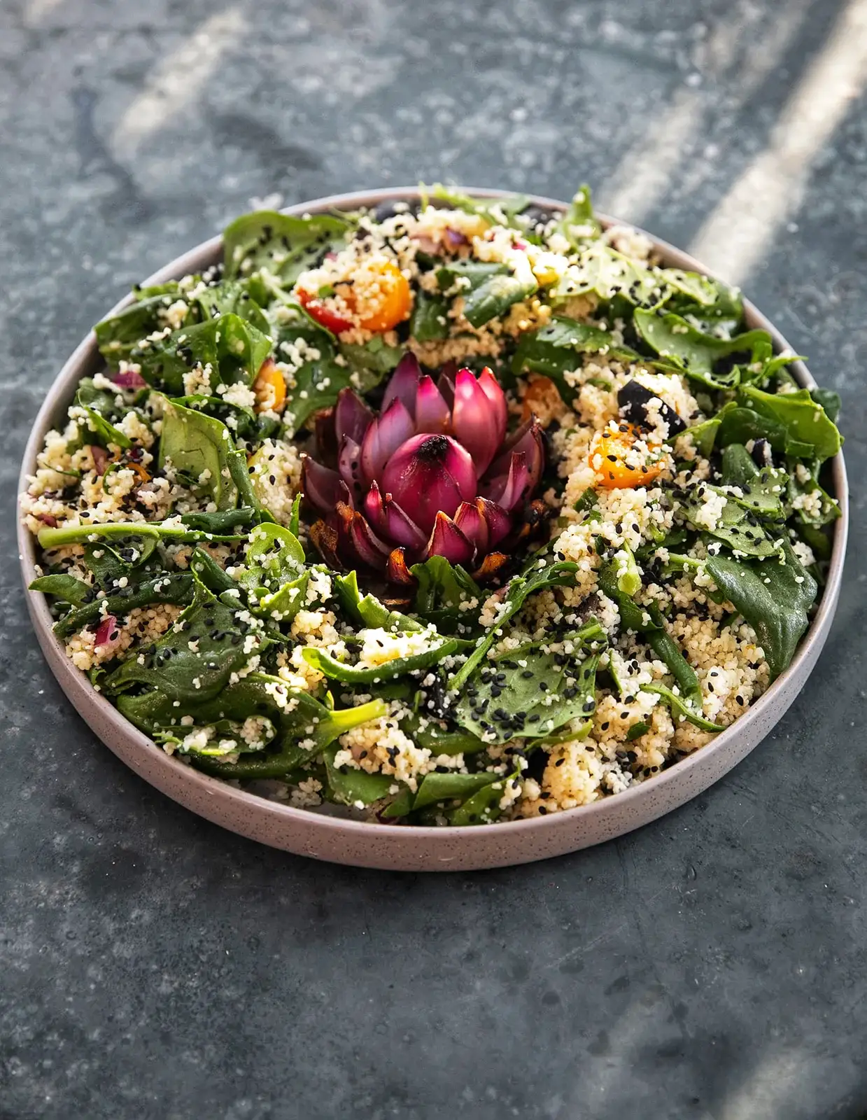 En färgstark sallad med bladgrönt, couscous, körsbärstomater, svarta frön och en lila blomma i mitten.