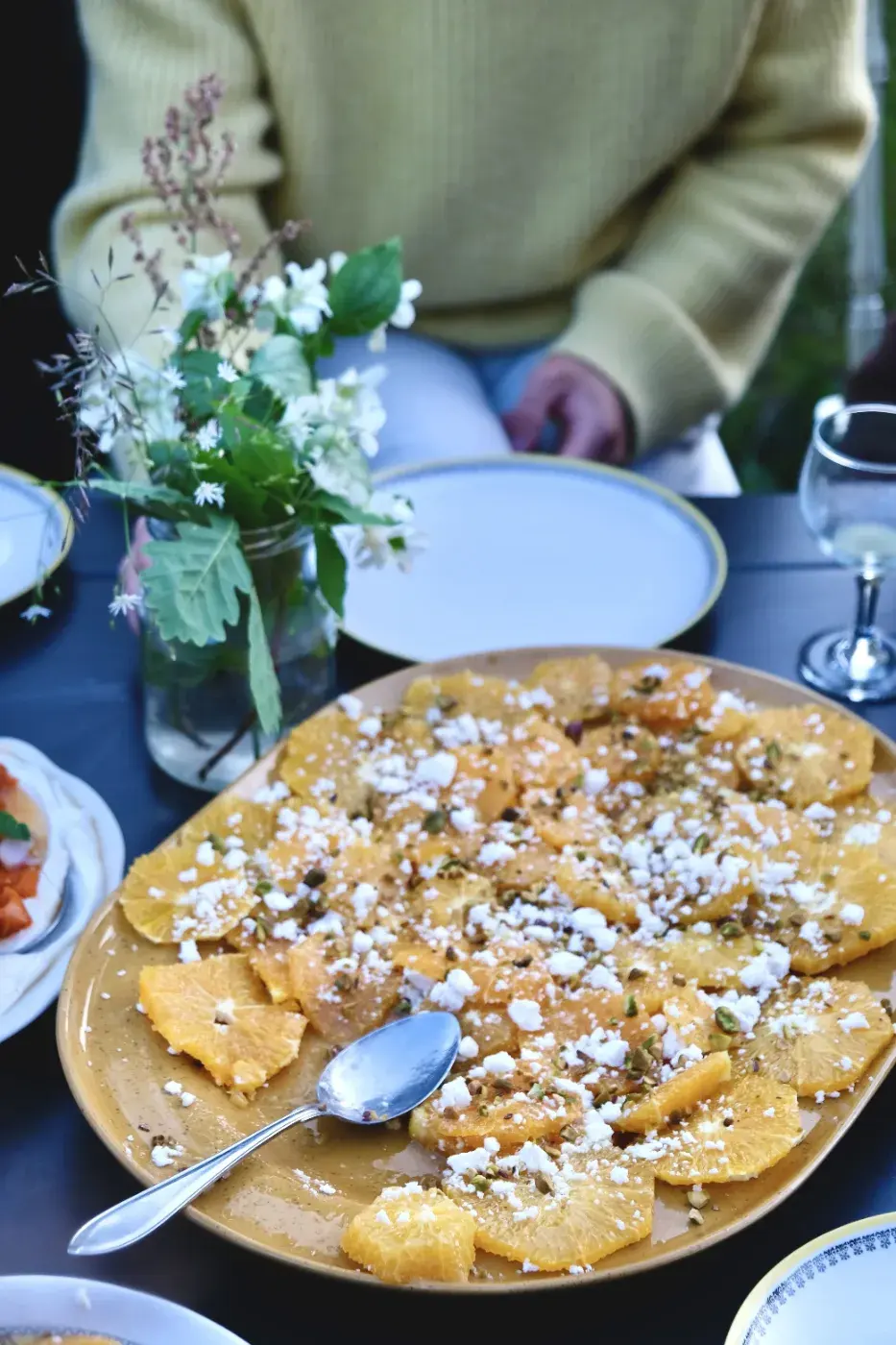 Ett upplagt fat med apelsinskivor, smulad ost och hackade nötter står på ett dukat bord med blommor i en vas och glas.