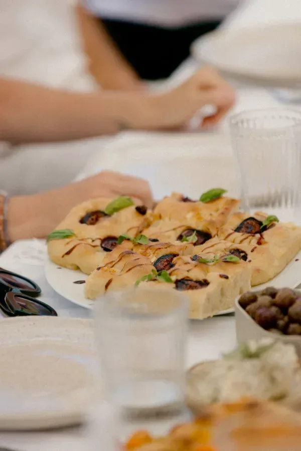 Focaccia med oliver och gröna örter ligger på ett fat på ett dukat bord, med personer i bakgrunden.