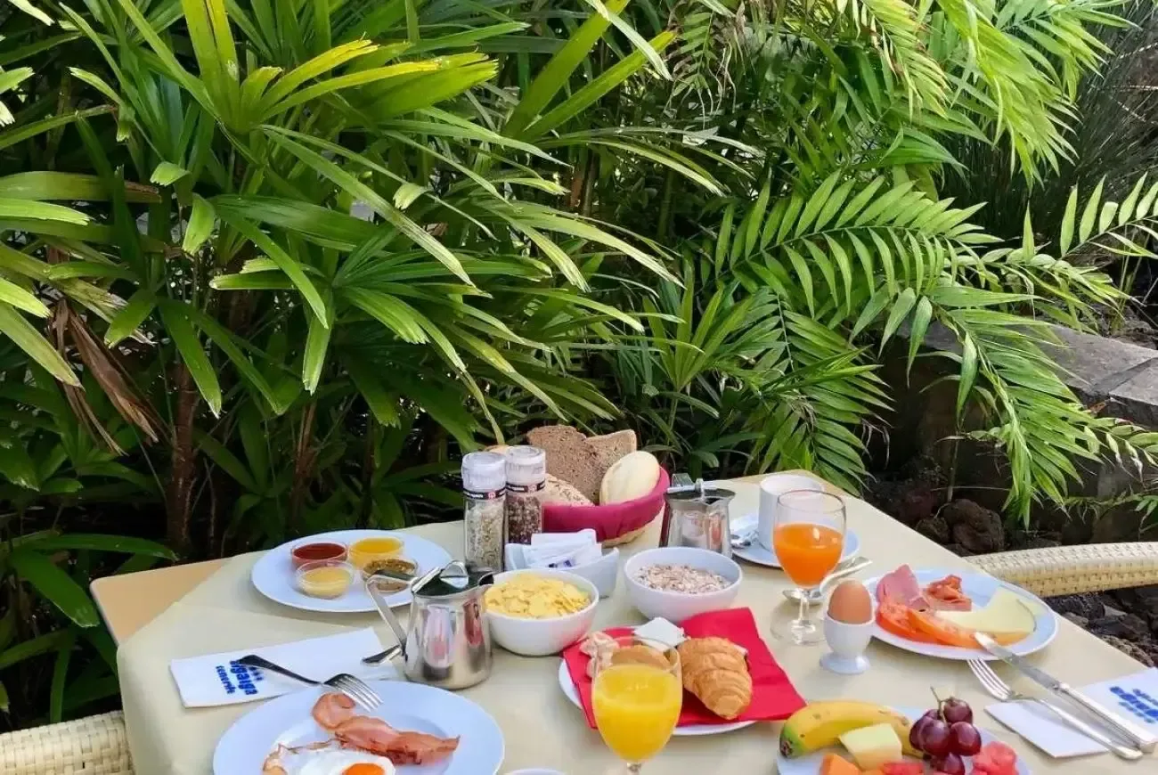 Ett dukat frukostbord med bröd, frukt, ägg, yoghurt, juice och kaffe omgivet av frodig tropisk grönska.