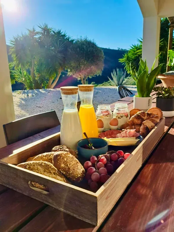 En frukostbricka med bröd, vindruvor, ägg, pålägg, yoghurt och juice står på ett bord utomhus med grönska och blå himmel i bakgrunden.