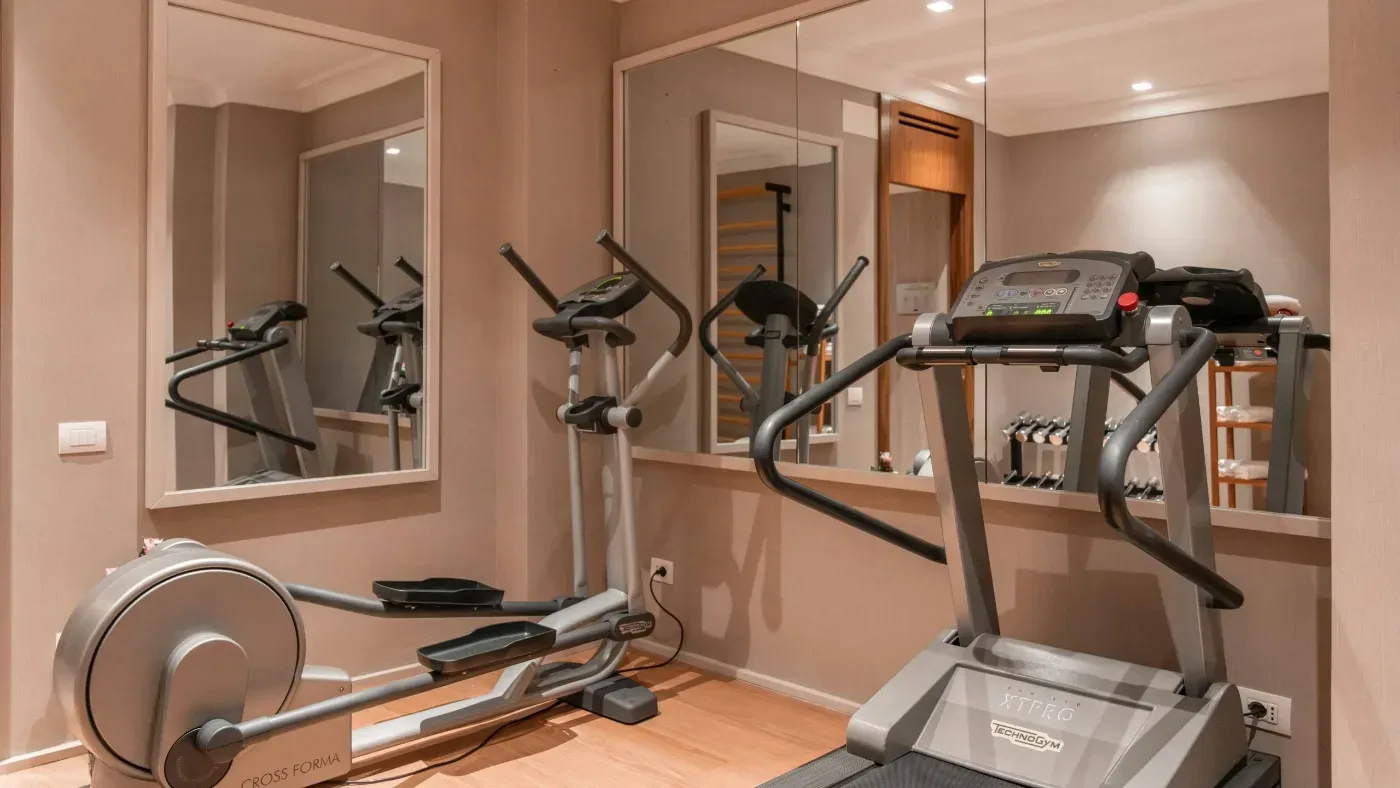 Ett litet gym med löpband, crosstrainer, motionscykel och hantlar framför spegelväggar.