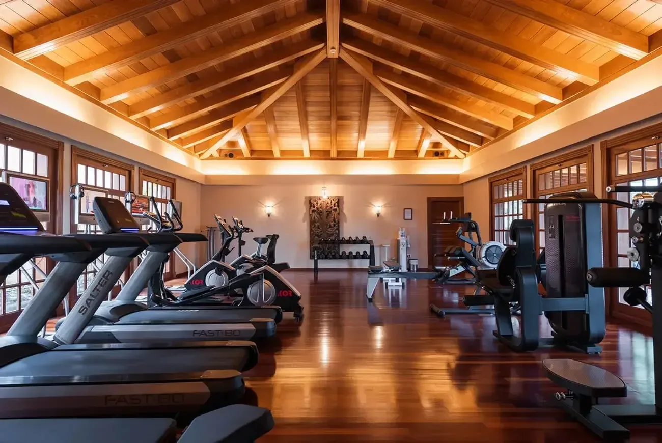 Ett ljust gym med träningsmaskiner, hantlar och roddmaskin i en rymlig lokal med synligt träbjälktak och stora fönster.