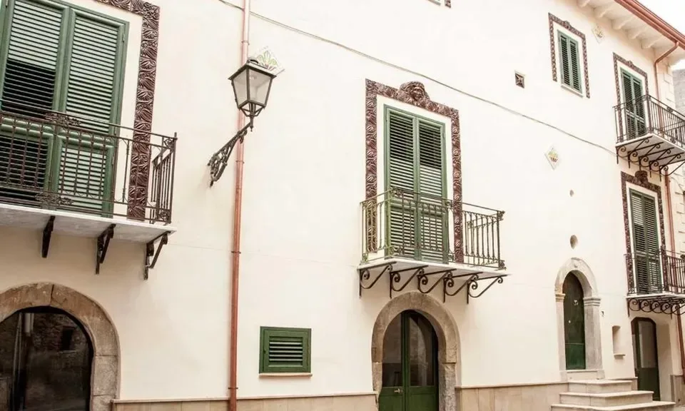 Ljus historisk byggnad i Santo Stefano di Camastra med gröna fönsterluckor, smidesbalkonger och välvda dörröppningar.