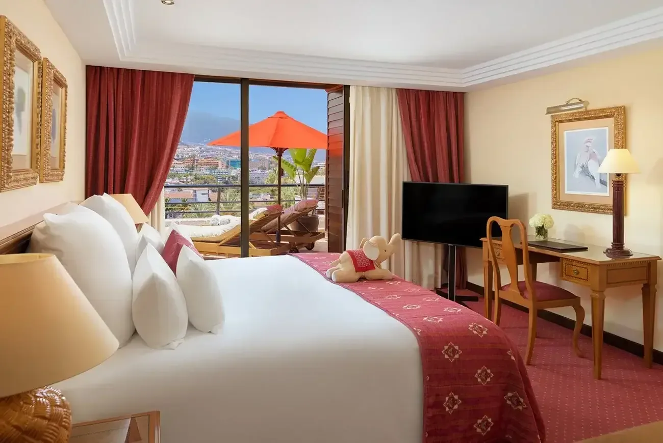 Elegant hotellrum med bäddad säng, skrivbord och balkong med utsikt över kuststaden och bergen på Teneriffa.