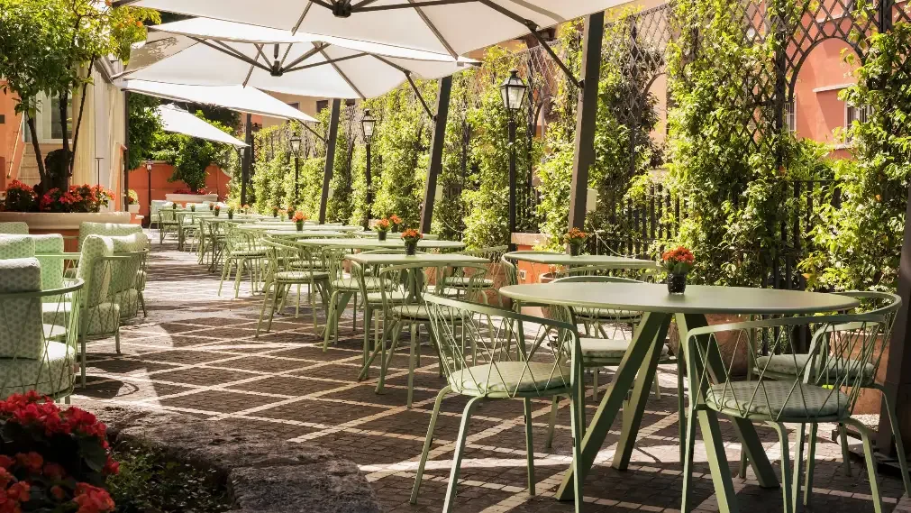 En solig innergårdsterrass i Rom med gröna cafébord, stolar, stora parasoller och blomkrukor omgiven av frodig växtlighet.