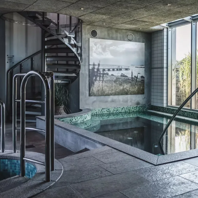 Inomhuspool med trappa, mosaikkant och stora fönster, samt ett svartvitt kustmotiv på väggen.