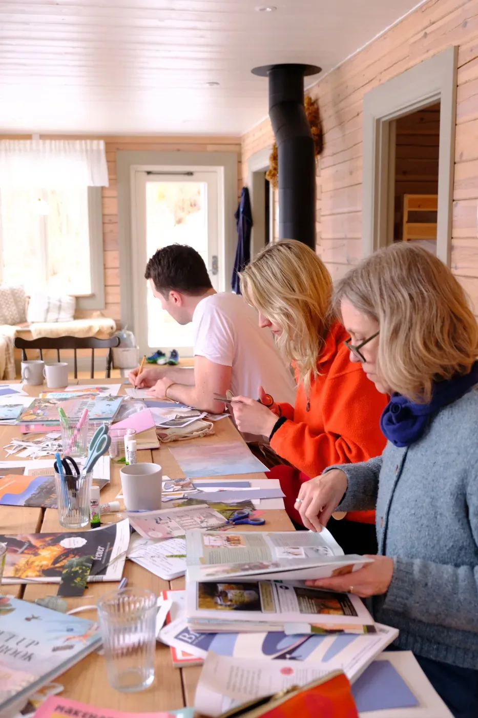 Tre personer arbetar kreativt vid ett bord med papper, böcker, saxar och annat material i en ljus trästuga med braskamin.