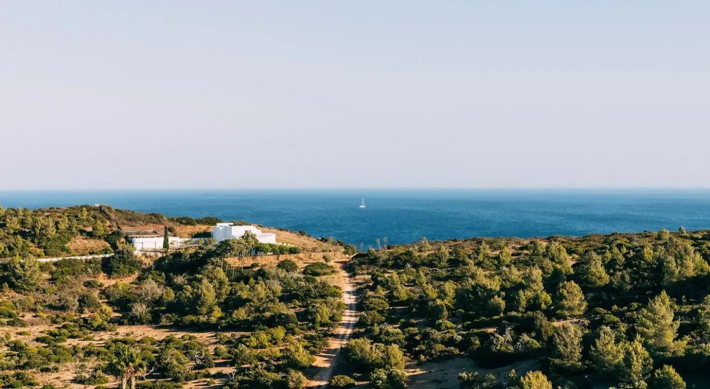 Ett kustlandskap i Algarve med en vit byggnad, en stig genom låg vegetation och Atlanten i bakgrunden.