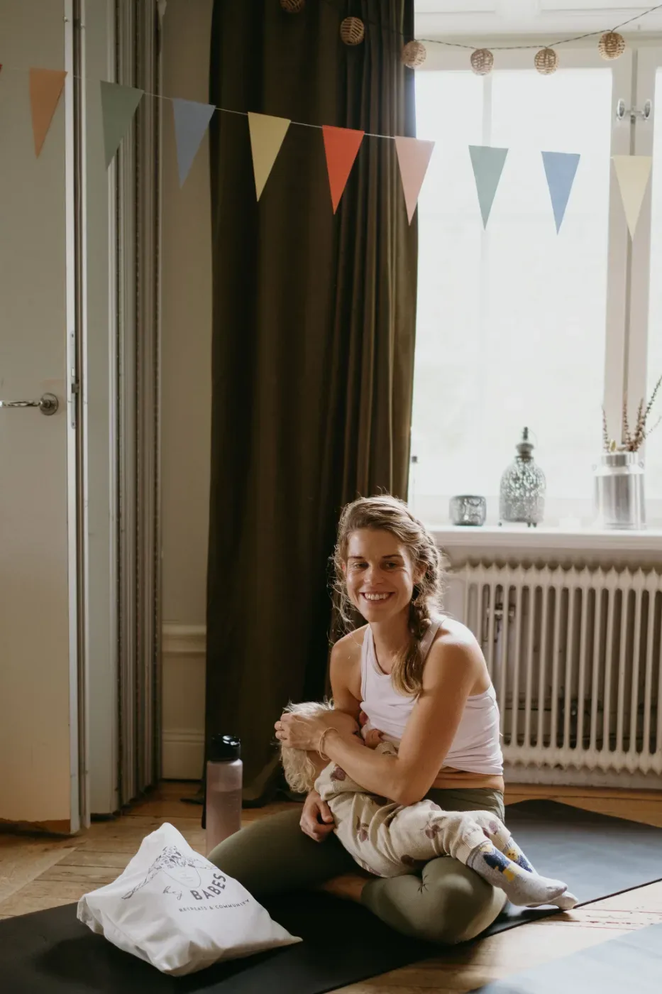 En leende kvinna sitter på en yogamatta med ett litet barn i famnen i ett ljust rum dekorerat med färgglada vimplar.