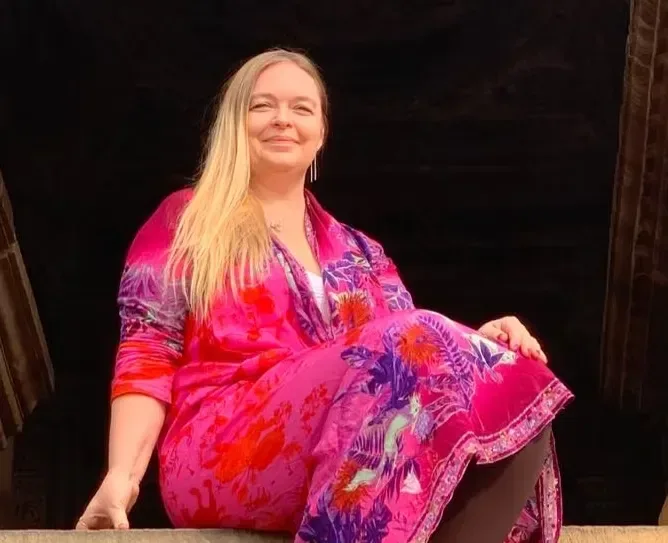 En leende kvinna med långt blont hår sitter i en dörröppning klädd i en färgstark rosa och lila blommig klänning.