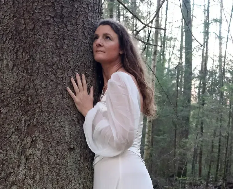 En kvinna i vit klänning står med händerna mot en stor trädstam i en skog.