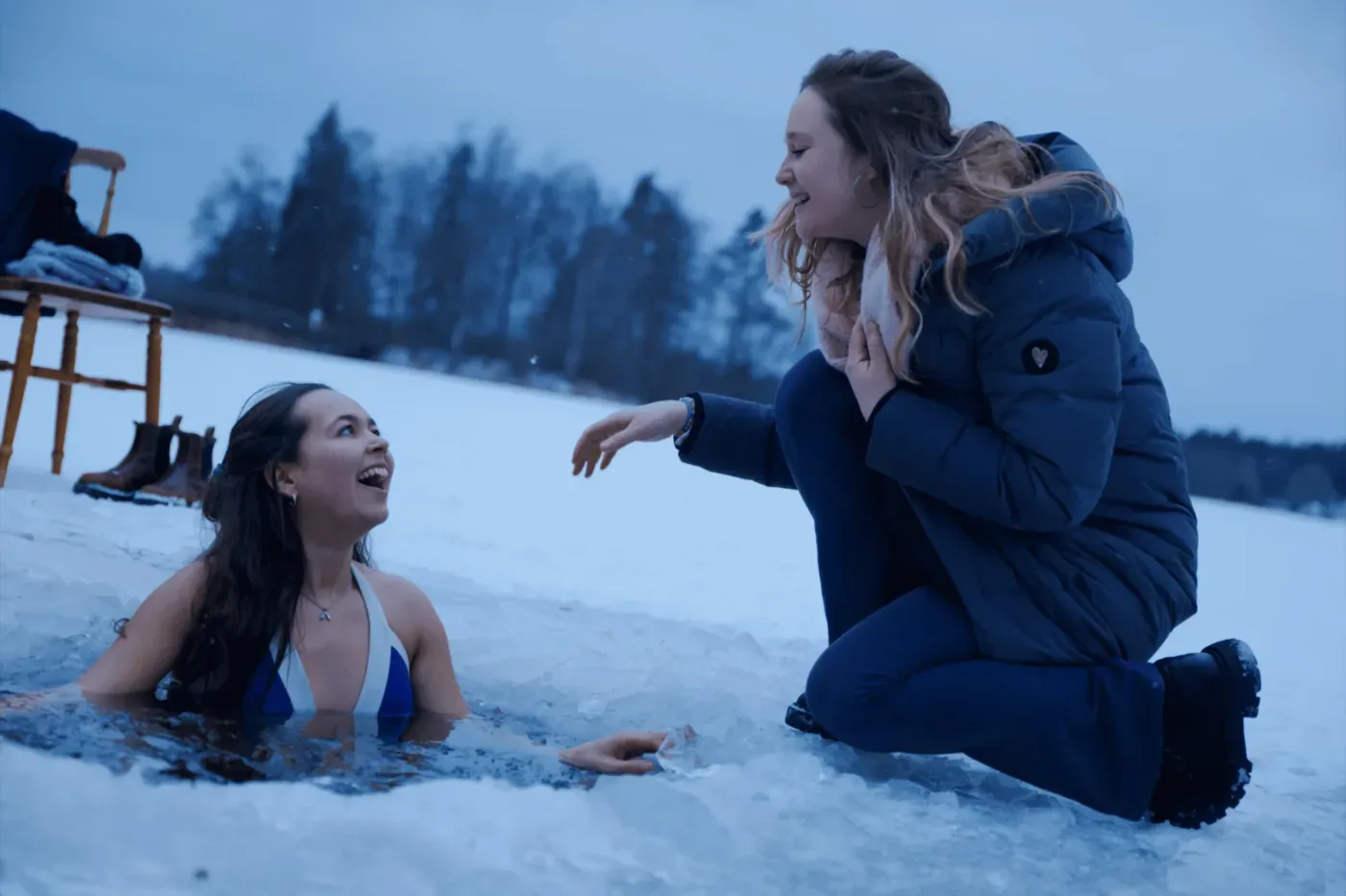 En kvinna sitter i en isvak medan en annan kvinna i vinterkläder knäböjer bredvid i det snötäckta landskapet.