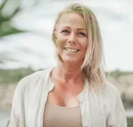 En leende kvinna med ljust hår står utomhus framför ett suddigt kustlandskap.