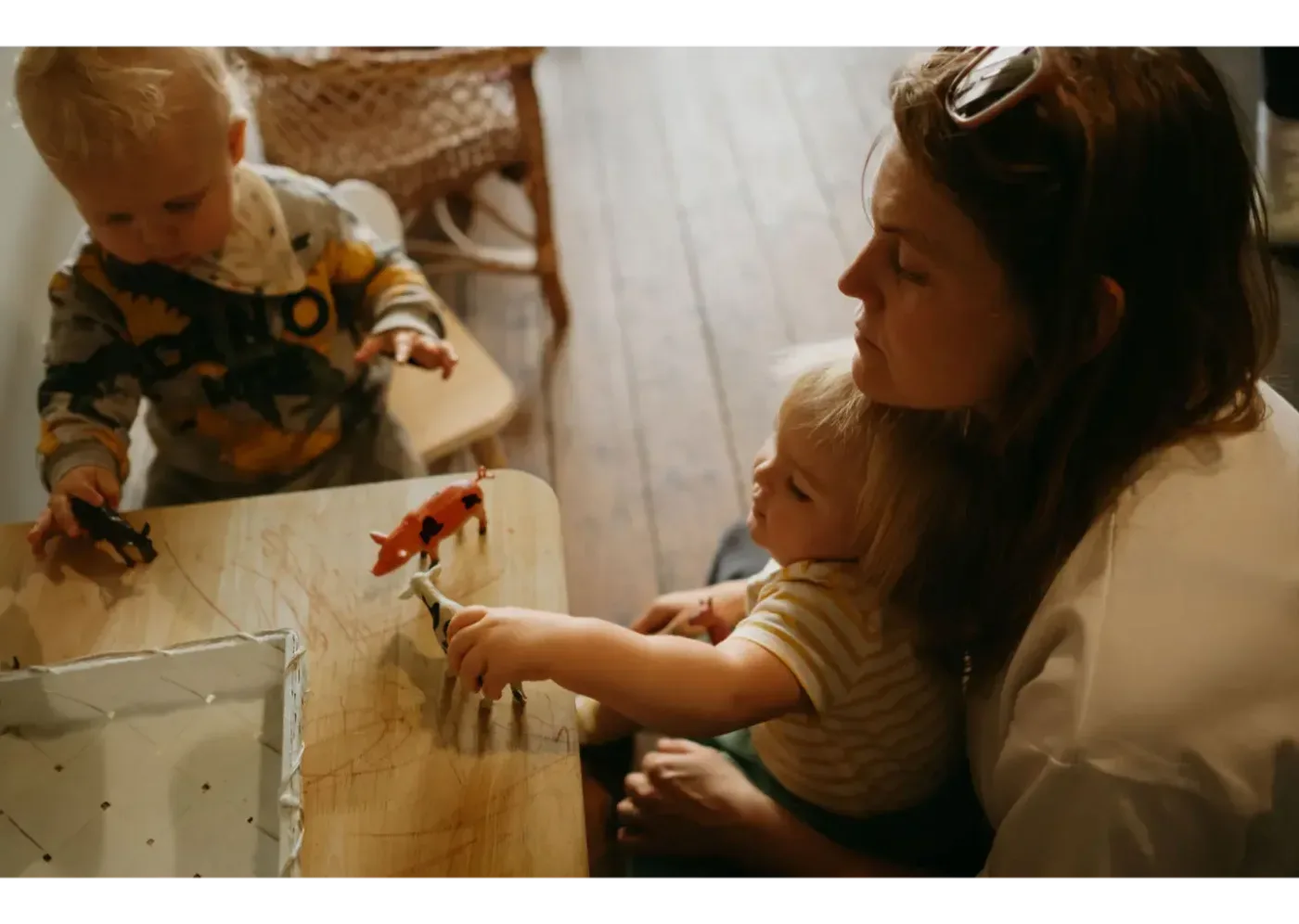 En mamma sitter bredvid två små barn som leker med leksaksdjur vid ett bord inomhus.