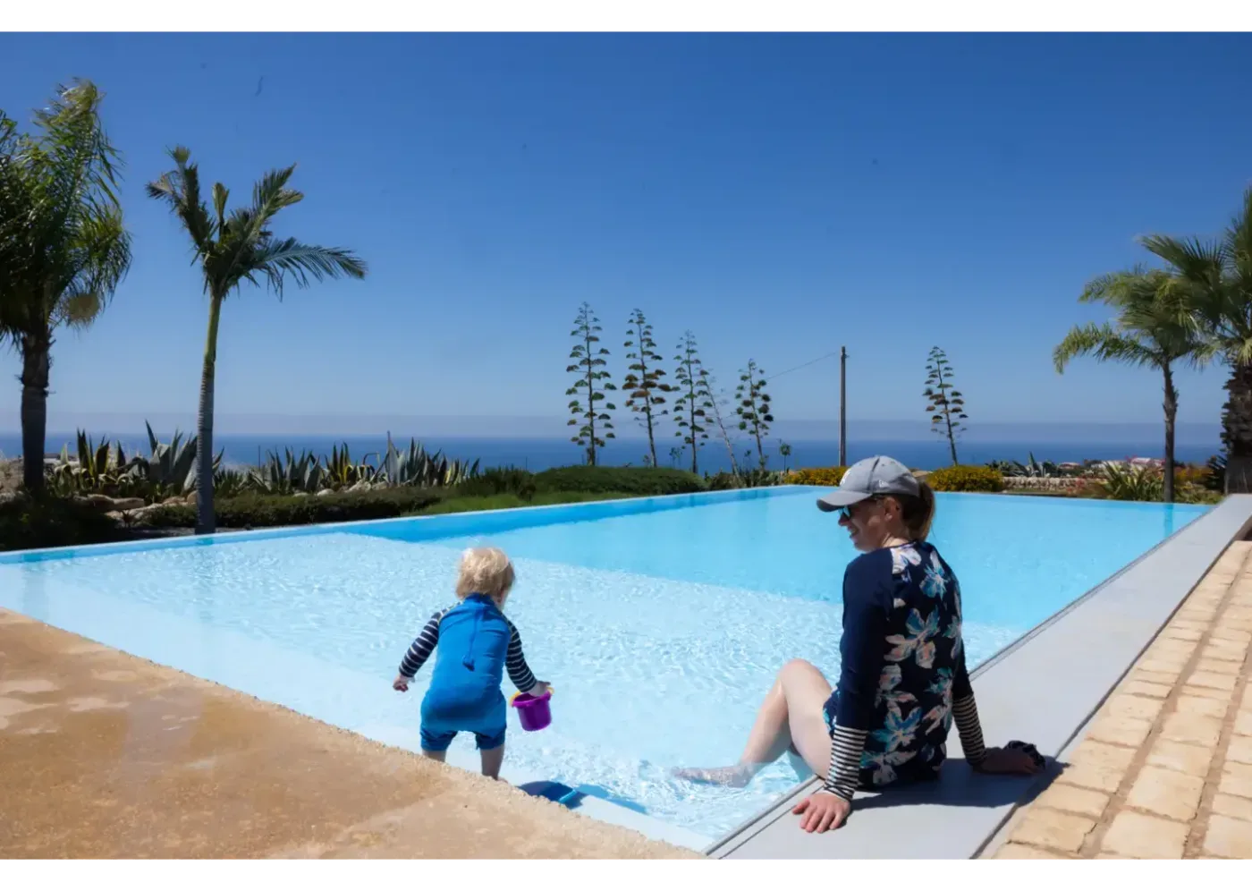 En kvinna sitter vid kanten av en pool medan ett barn leker i vattnet med en hink, med havet och palmer i bakgrunden.