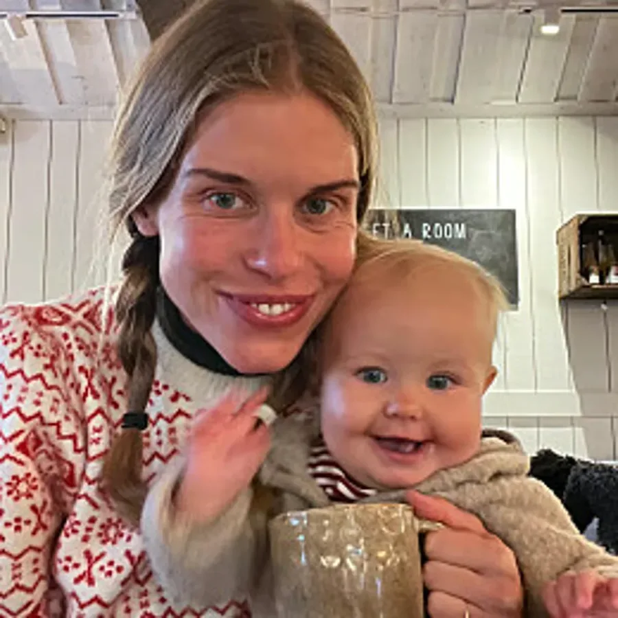 En kvinna håller ett leende spädbarn i famnen inomhus, med en mugg i förgrunden.