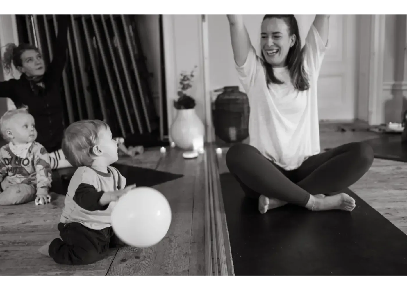 En kvinna sitter på en yogamatta med armarna sträckta uppåt medan två små barn leker på golvet med en ballong och en annan vuxen sitter bakom dem.