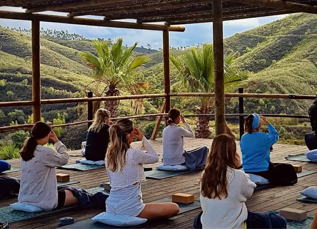 En grupp deltagare sitter på yogamattor under ett trädäck och utövar meditation med utsikt över gröna kullar och palmer i Algarve.