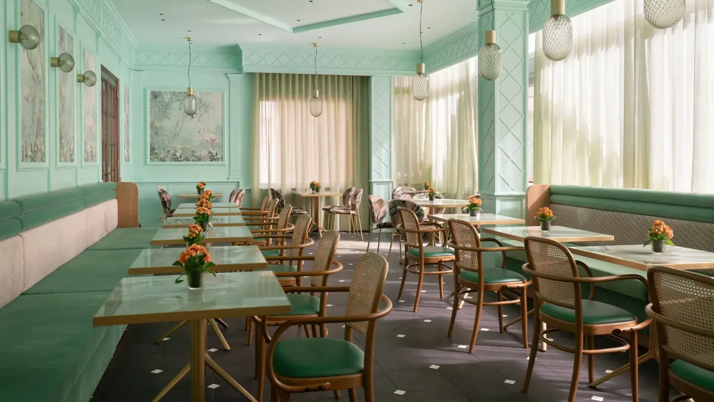 Ljust mintgrönt café med dukade bord, gröna soffor, rottingstolar och små blomvaser vid stora fönster.