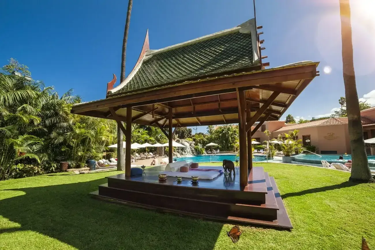 En öppen paviljong med madrasser och kuddar står i en grönskande trädgård intill en pool under palmer och klarblå himmel.