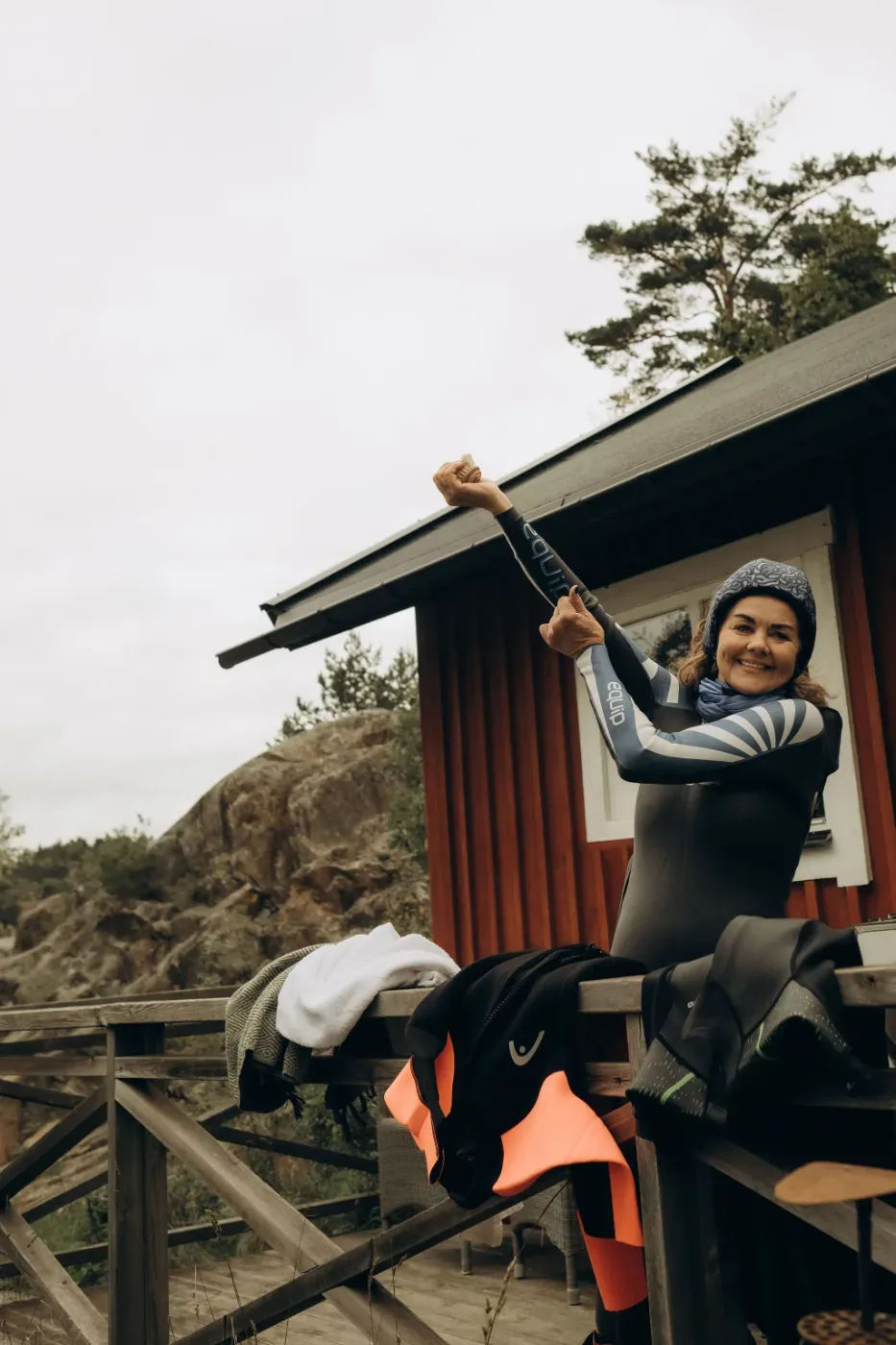 En person i våtdräkt sträcker armarna framför en röd trästuga, med klippor och tallar i bakgrunden.