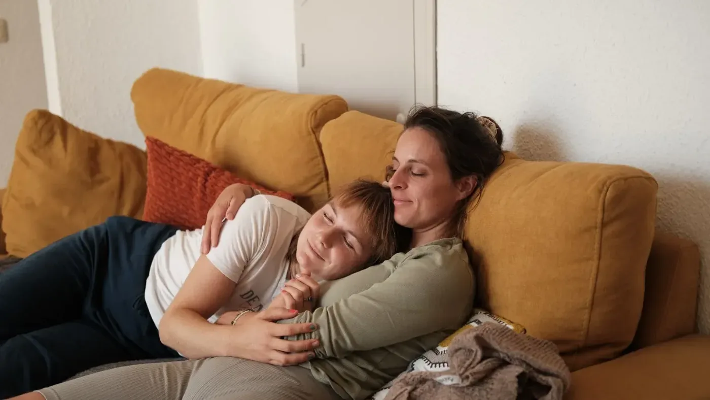 Två personer sitter tätt tillsammans i en senapsgul soffa, där den ena vilar med huvudet mot den andra.