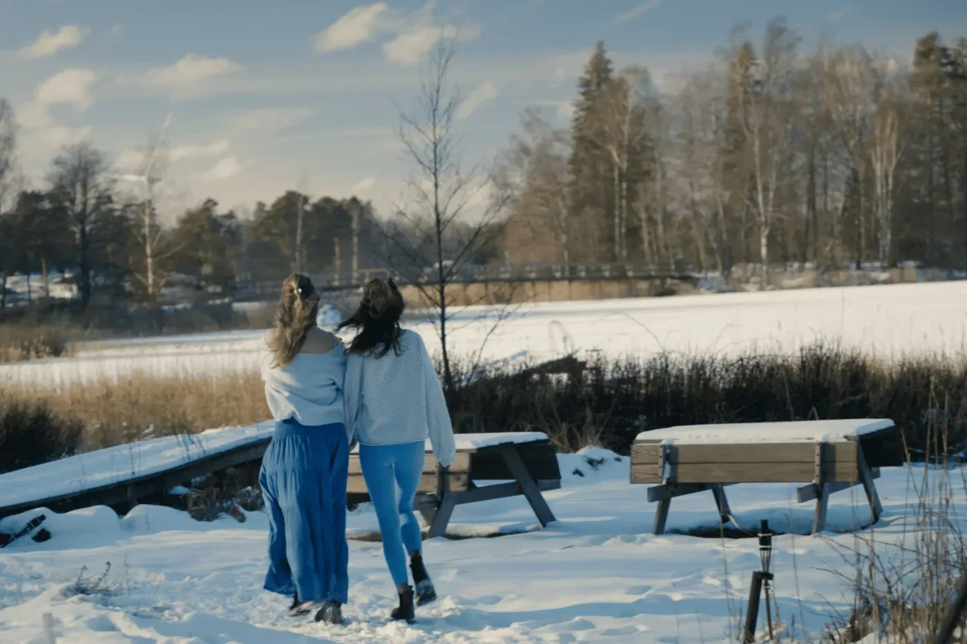 Två personer går tillsammans genom ett snötäckt landskap med träbänkar, en bro och skog i bakgrunden.