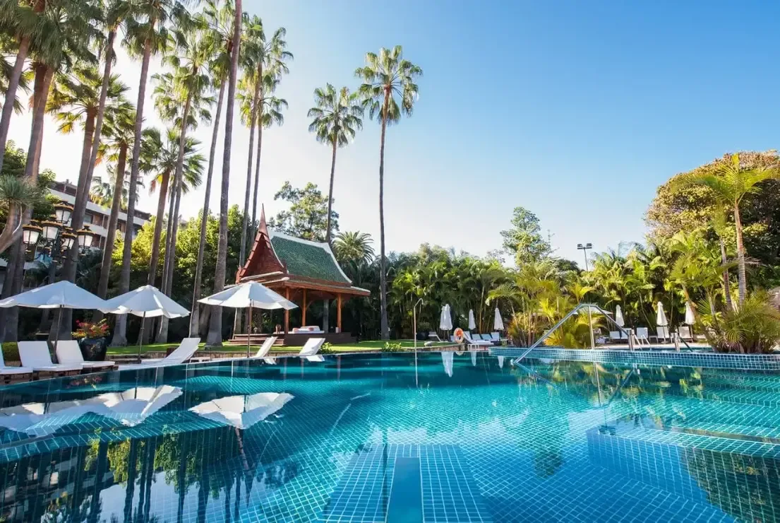 En stor pool omgiven av palmer, solstolar och vita parasoller, med en thailändskinspirerad paviljong i den lummiga trädgården.