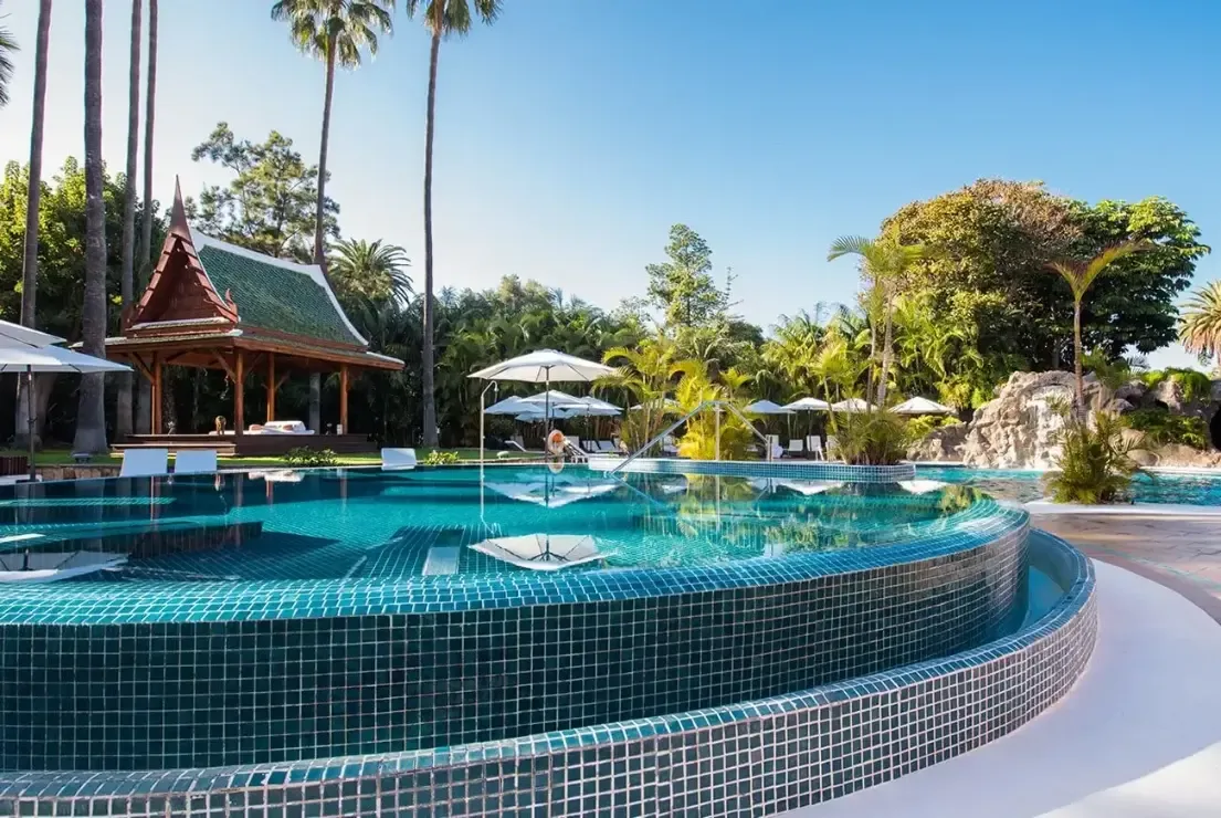 En stor mosaikklädd pool omgiven av palmer, parasoller och en thailändskinspirerad paviljong.