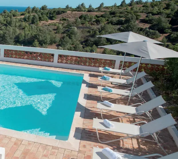 En turkos pool omgiven av solstolar och parasoller, med ett kuperat landskap och havet i bakgrunden.