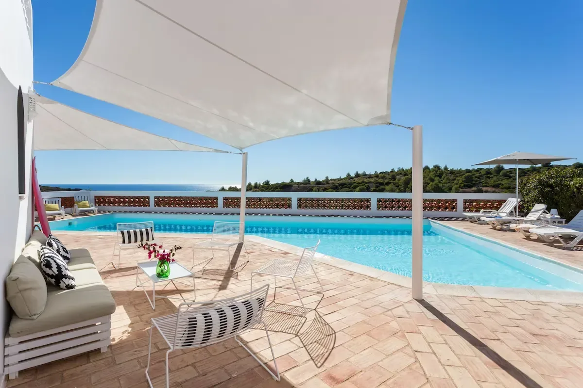 Poolområde med solstolar, sittgrupp och skuggande segelduk med utsikt över havet i Algarve.