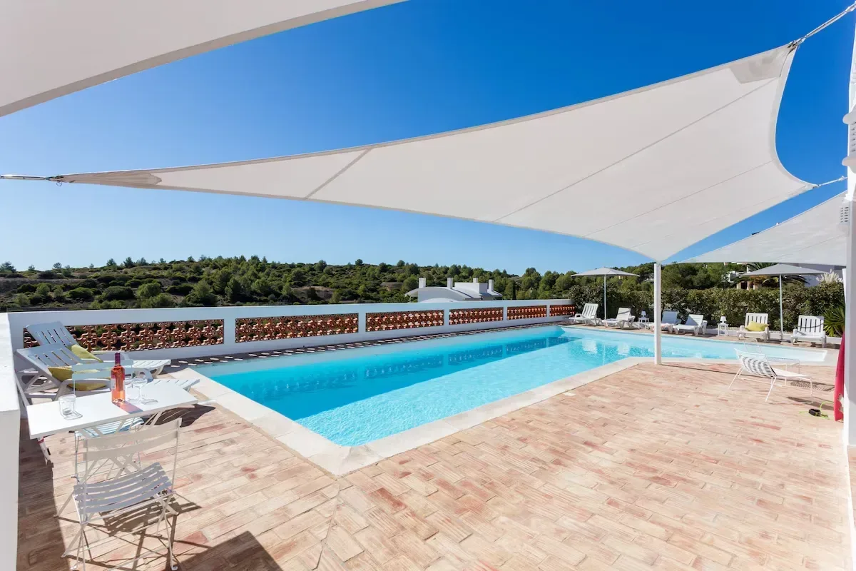 En solig terrass med en turkos pool, vita solstolar och stora segeldukar framför ett grönt landskap i Algarve.