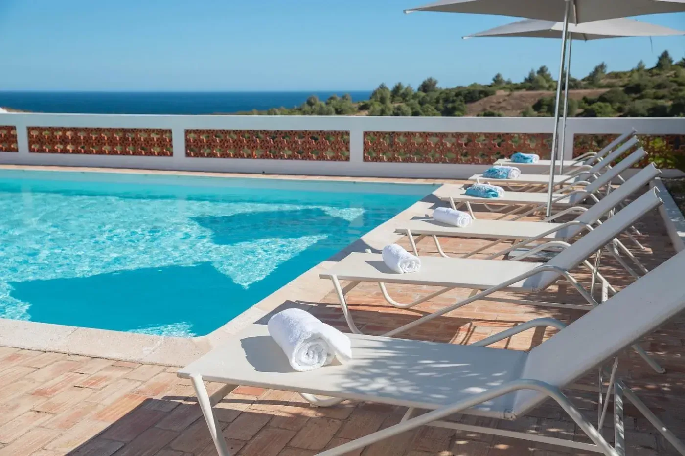 En solig poolterrass med vita solstolar, hoprullade handdukar och utsikt över havet och det gröna landskapet i Algarve.