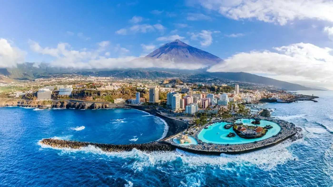 Vy över Puerto de la Cruz på Teneriffa med kust, havsbassänger, stadsbebyggelse och Teide i bakgrunden.